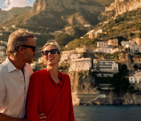 A couple of guests sails on a leisure boat not far from the Amalfi Coast. They look at each other and smile, enjoying the moment. He embraces her tenderly.