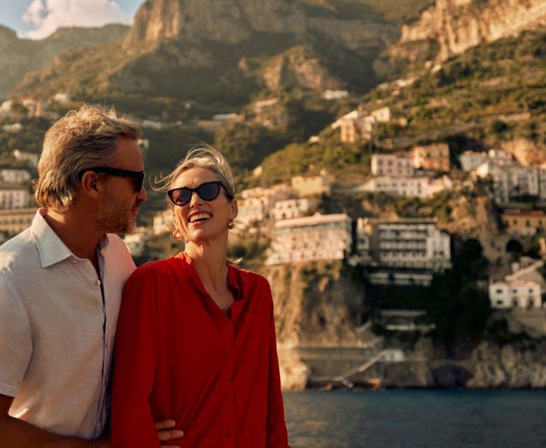 A couple of guests sails on a leisure boat not far from the Amalfi Coast. They look at each other and smile, enjoying the moment. He embraces her tenderly.